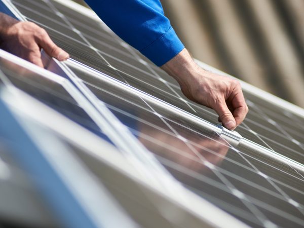 Male engineer in blue suit installing solar photovoltaic panel system. Close up view of electrician hands mounting blue solar module on roof of modern